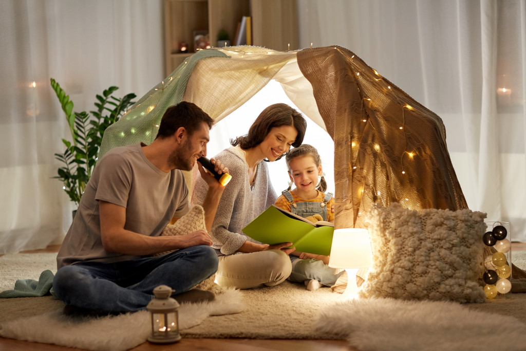 A family of three reading a book in an indoor tent made of blankets, pillows, and string lights